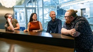 Four colleagues sit at a wooden table in a modern office lobby, smiling and chatting, with glass walls, staircase, and natural light in the background.