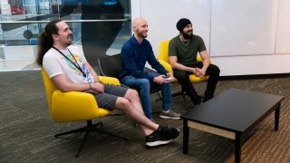 Three men sit in colorful lounge chairs in a modern office, smiling and playing a video game with a controller.