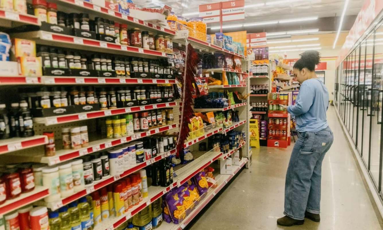 A person stands in a grocery aisle, browsing a variety of spices, sauces, and snacks on the shelves.