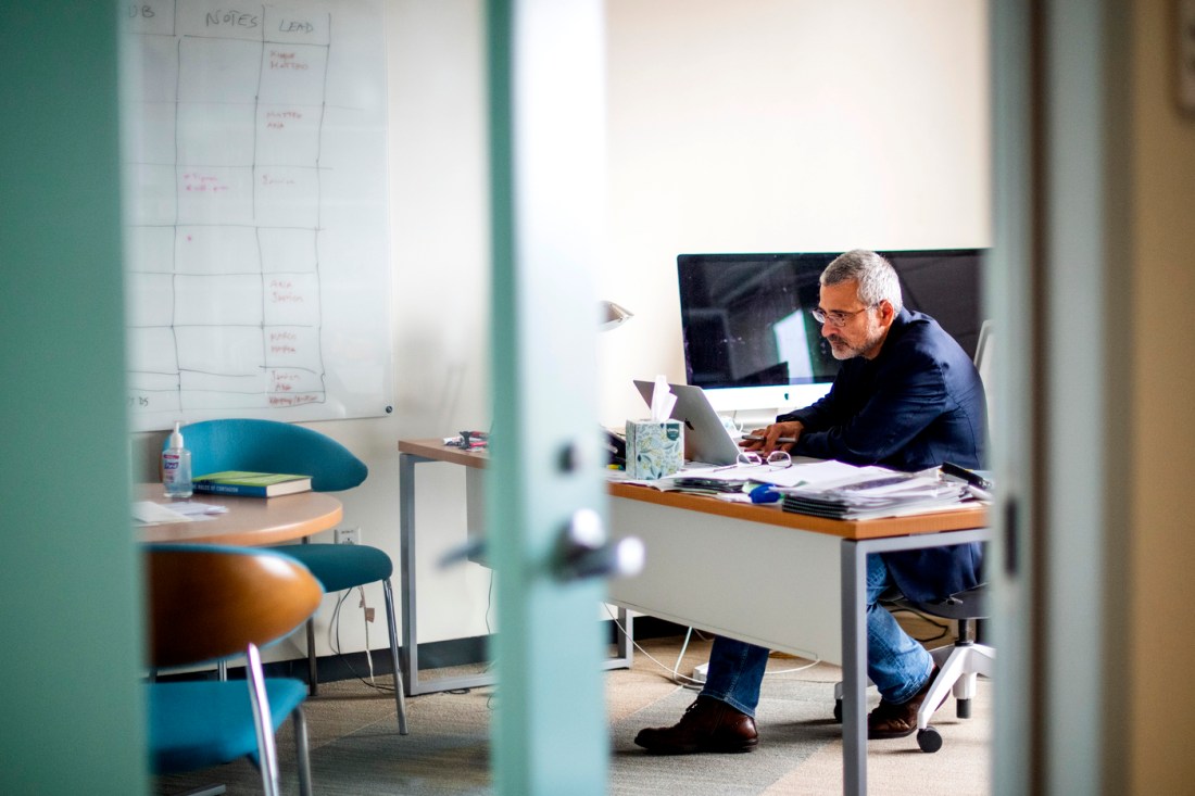 Alessandro Vespignani sitting at a desk working at a laptop inside of an office. In the foreground, the open door into the office is blurred.
