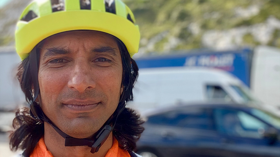 Vijay, wearing a yellow helmet and orange cycling jacket, snaps a selfie beside a hilly background