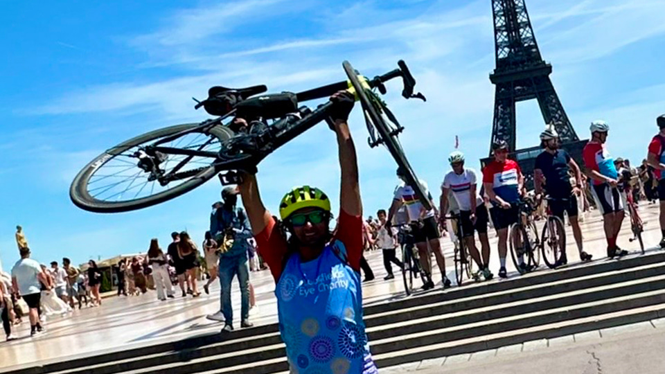 Vijay holds his bike above his head in front of the Eiffel Tower on a blue-sky sunny day