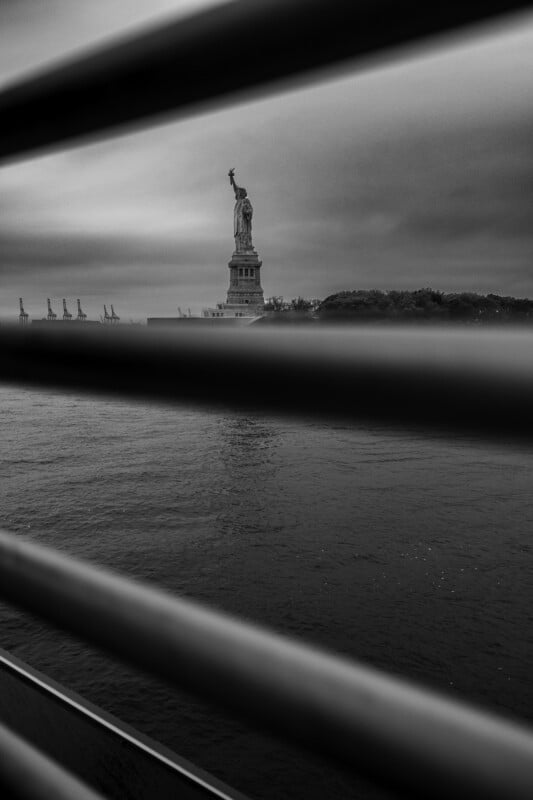 The Statue of Liberty is seen in the distance across the water, framed by horizontal metal bars in the foreground. The image is black and white, with a cloudy sky above.