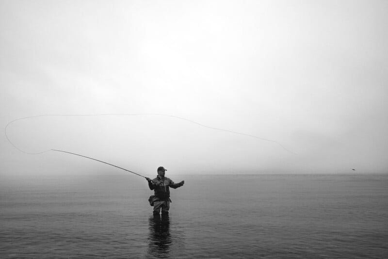 A person stands waist-deep in calm water, fly fishing under a cloudy sky. The fishing line arcs gracefully above them, and mist obscures the distant horizon. The scene is in black and white.