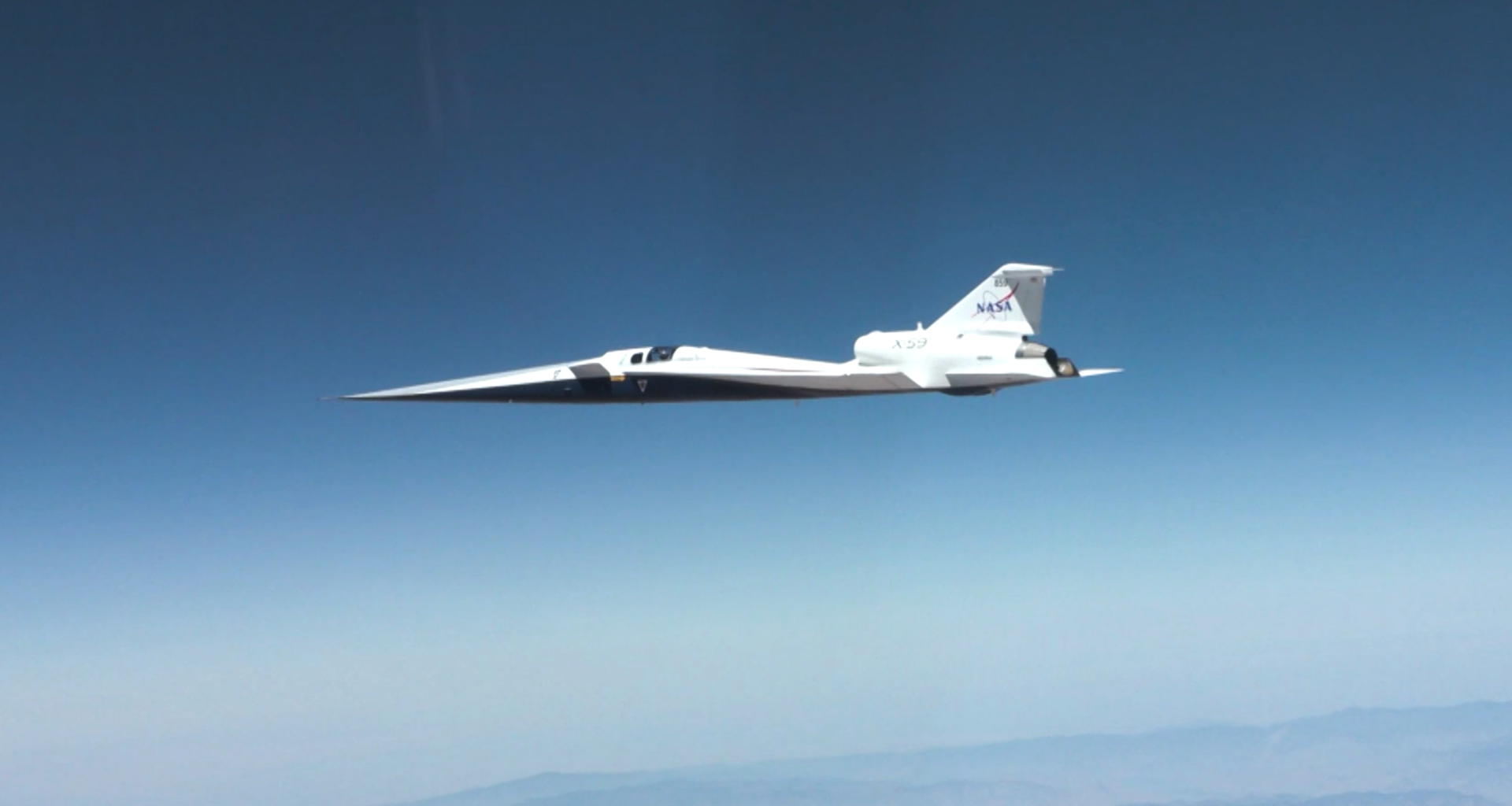 NASA’s X-59 quiet supersonic research aircraft flies through the blue sky. The plane has a distinctive long, sharp nose. There are mountains visible at the bottom of the photo.