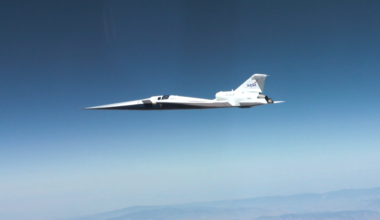 NASA’s X-59 quiet supersonic research aircraft flies through the blue sky. The plane has a distinctive long, sharp nose. There are mountains visible at the bottom of the photo.