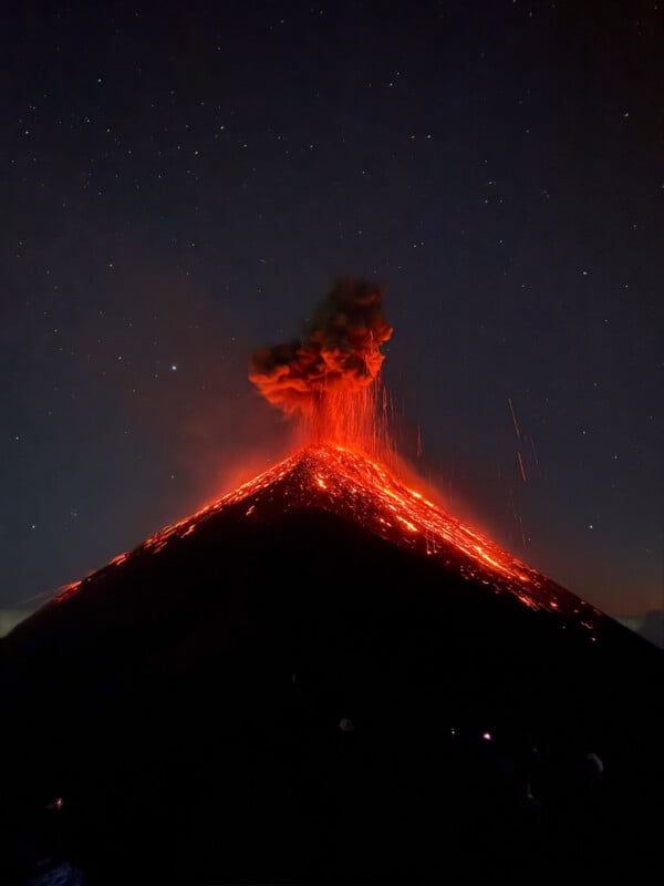 A volcano erupts at night, spewing bright red lava and smoke under a starry sky. Glowing lava streams flow down the dark slopes, illuminating the eruption.