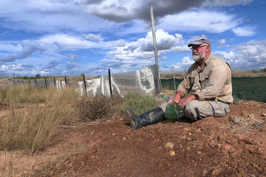 A man with gumboots on sits near a long shiny metal fence with mesh dividing a small puddle of water.