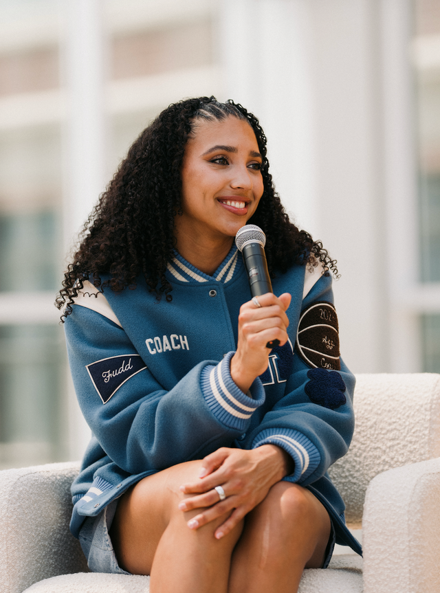 Smiling person with curly hair wearing a blue varsity jacket, holding a microphone while seated.