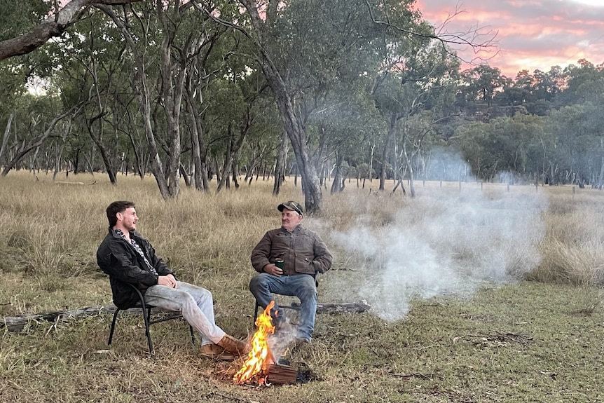 Two men sit in campchairs beside a fire at a rural property. The sun is setting behind them in shades of pink.