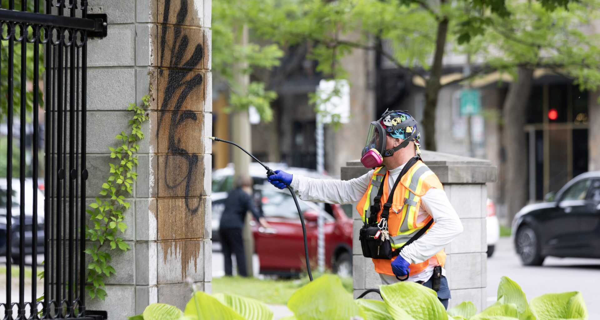 A man in an orange safety vest and safety mask pressure-washes graffiti from a cement column.