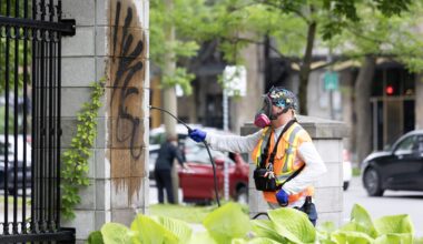 A man in an orange safety vest and safety mask pressure-washes graffiti from a cement column.