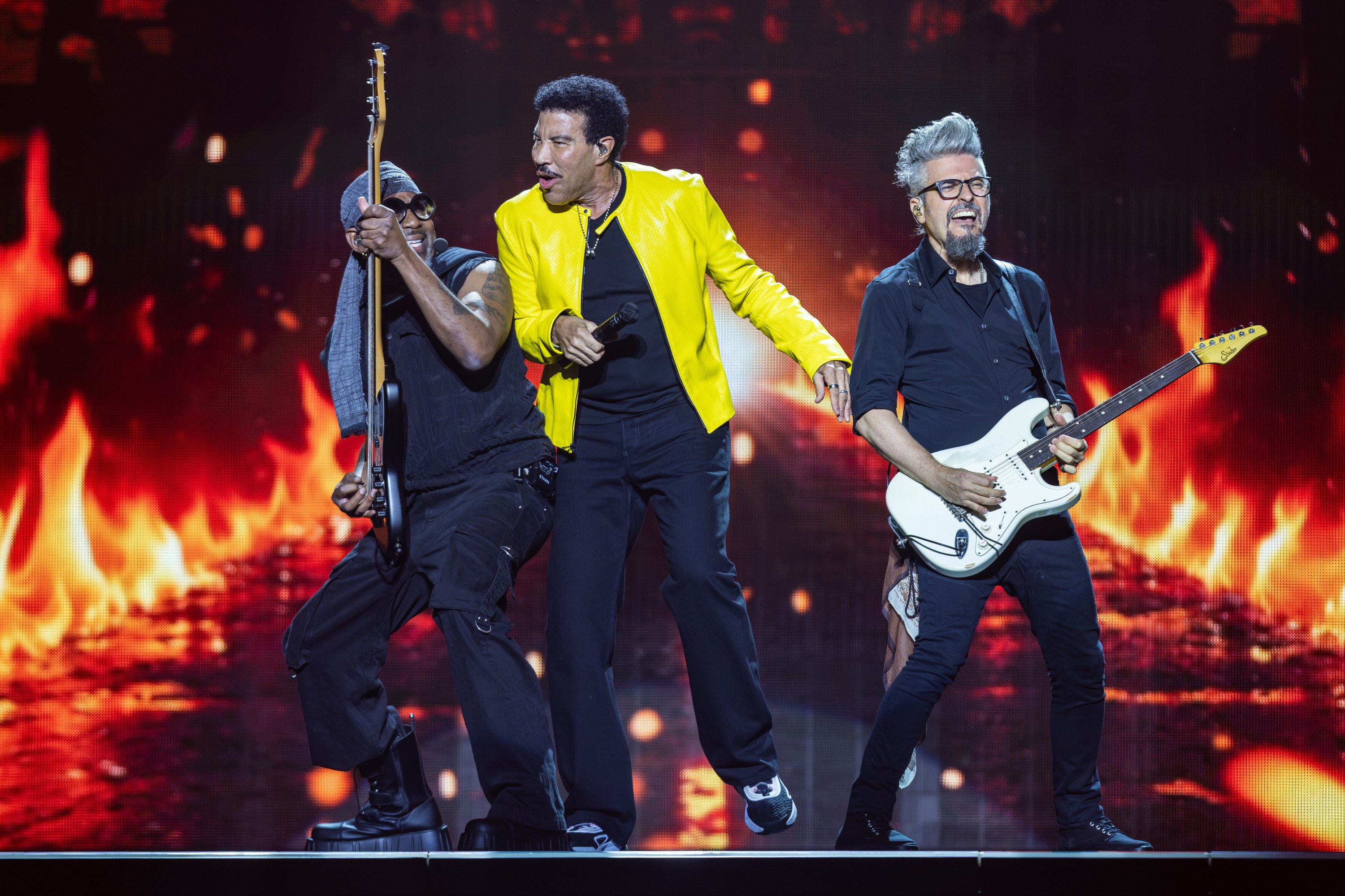 Lionel Richie, in a yellow leather jacket, moving in sync with guitarists flanking him.