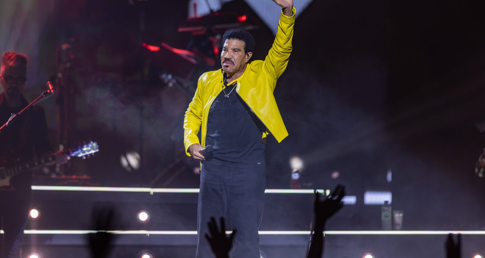 Lionel Richie waves to a crowd while on stage wearing a yellow jacket