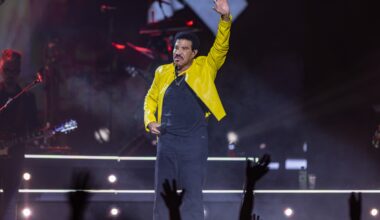 Lionel Richie waves to a crowd while on stage wearing a yellow jacket