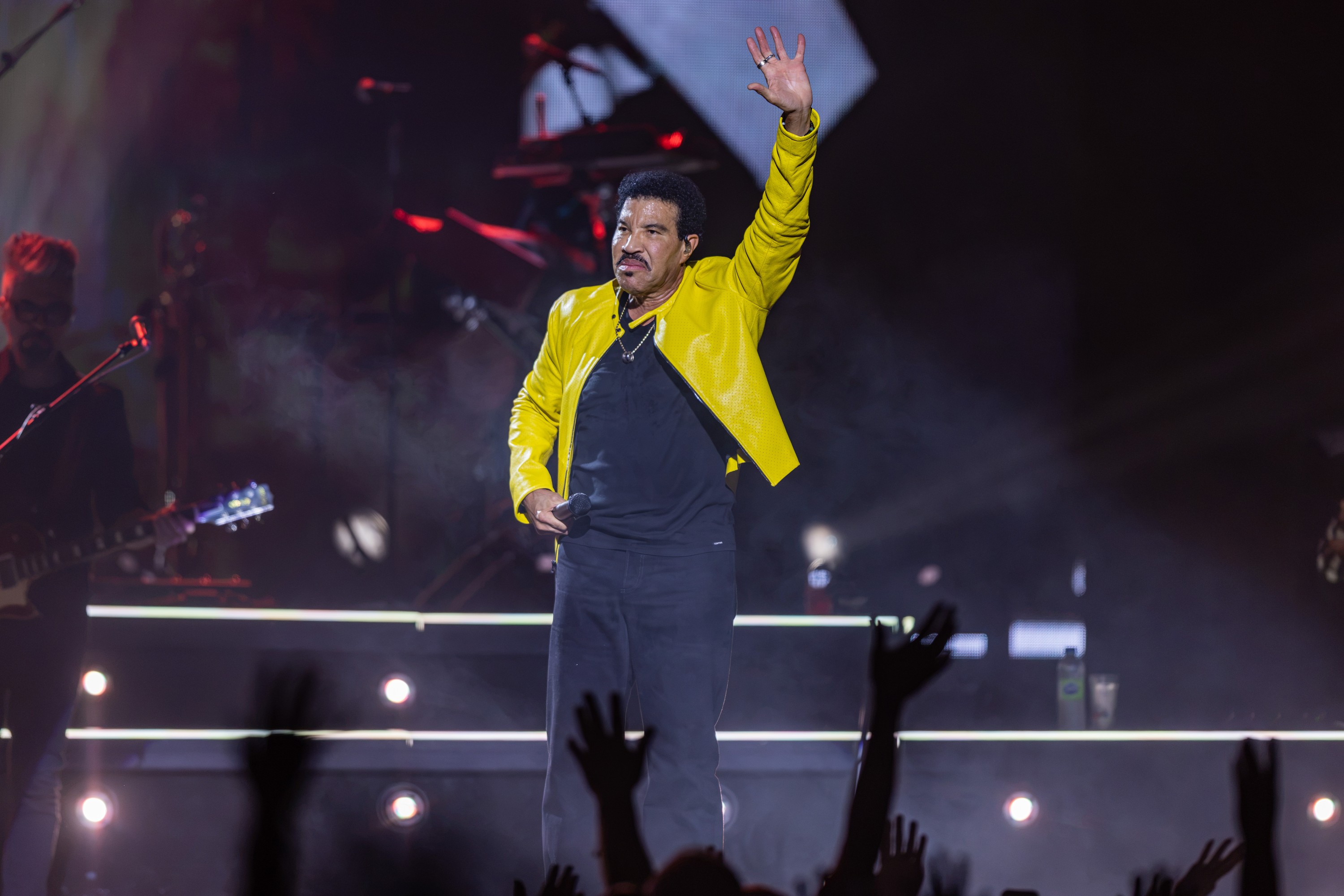 Lionel Richie waves to a crowd while on stage wearing a yellow jacket