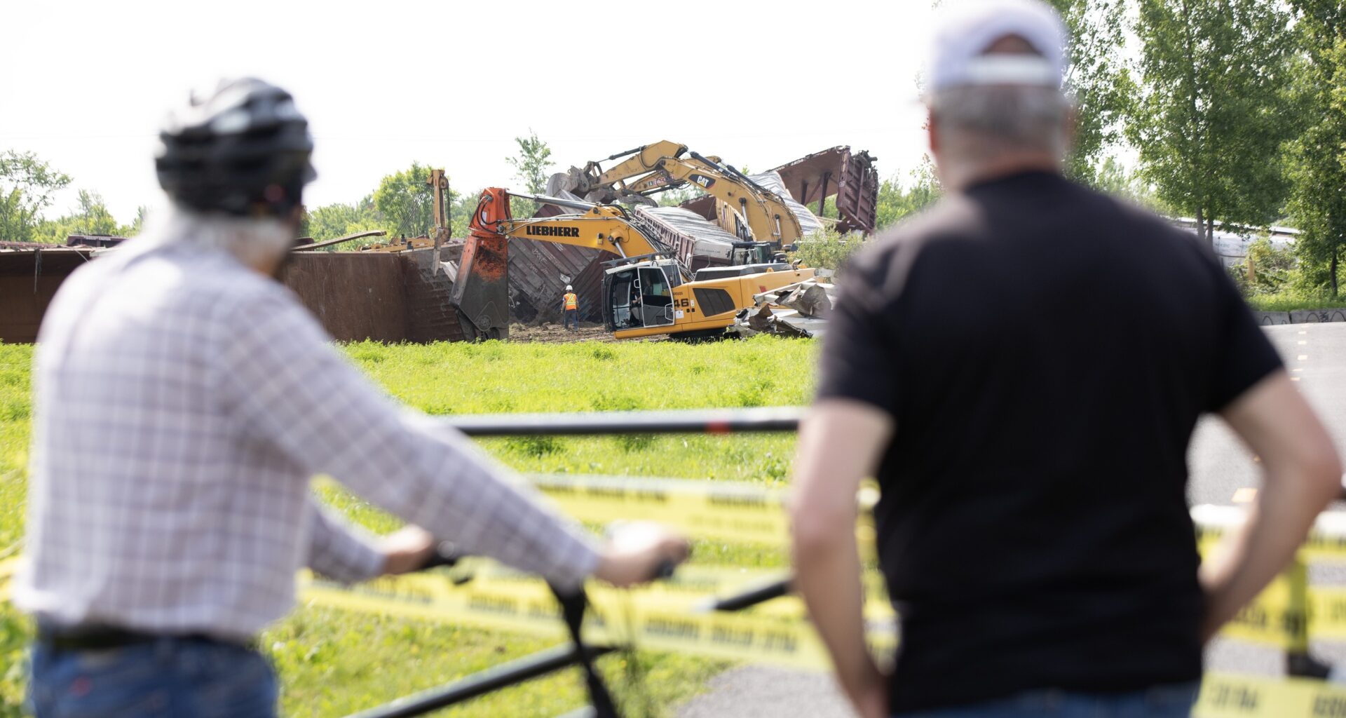 Two men stand behind caution tape as workers work to clean up after a train derailment.