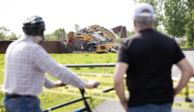 Two men stand behind caution tape as workers work to clean up after a train derailment.