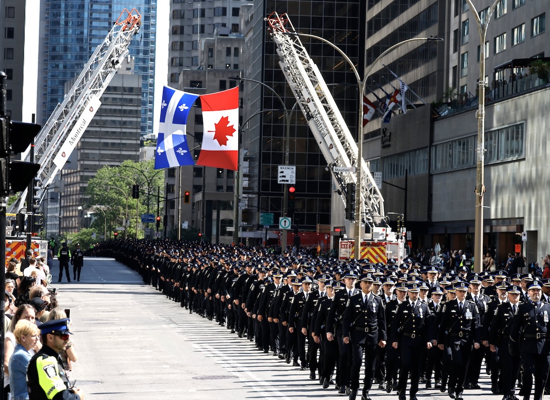 Hundreds of police officers walk in procession along a city street. Two fire-truck ladders are holding up the Canadian and Quebec flags. Citizens watch from the sidewalk.