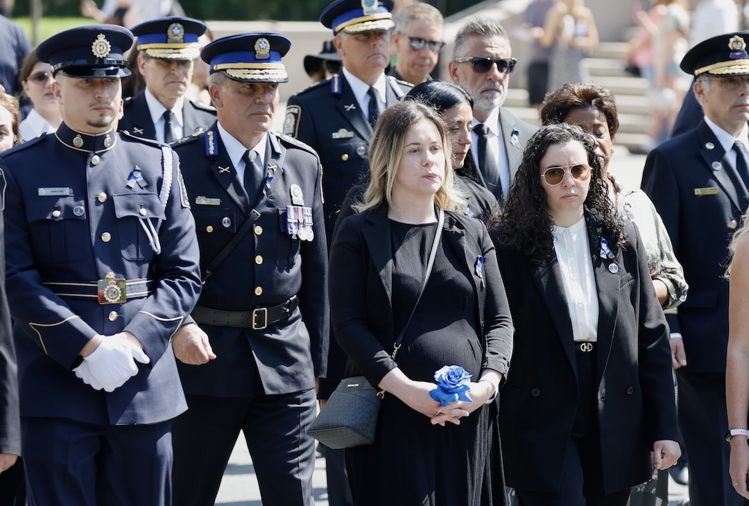 A pregnant woman wearing black and holding a blue rose stands with a woman and several police officers in dress uniforms.
