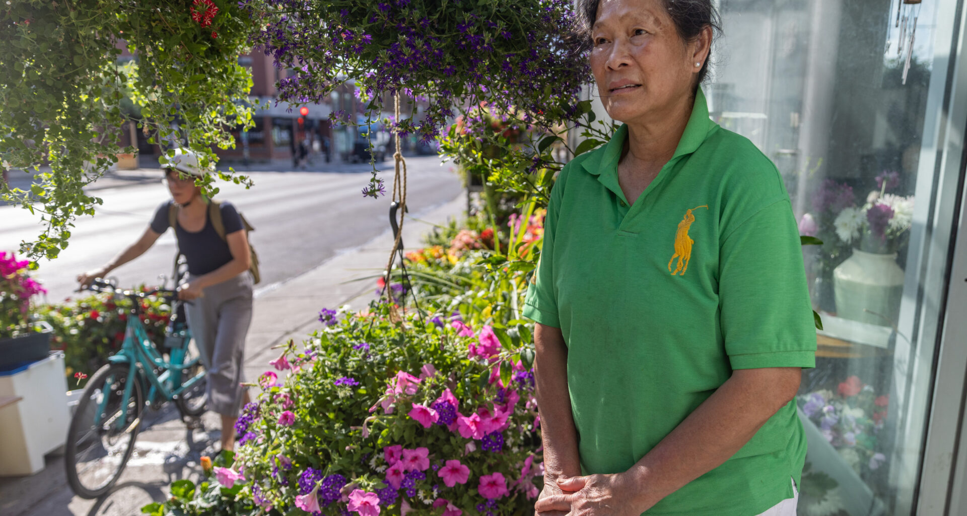A woman stands outside a flower shop as a person on a bicycle cycles by.