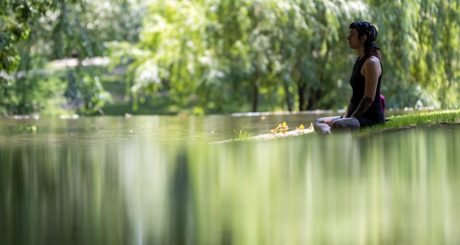 A woman sits on the edge of a pond, cross-legged with her arms at her sides and her eyes closed.