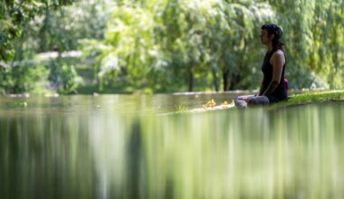 A woman sits on the edge of a pond, cross-legged with her arms at her sides and her eyes closed.