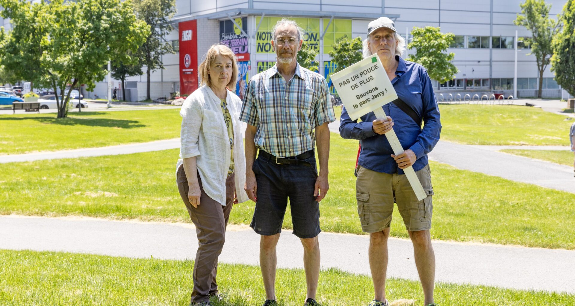Three people stand on a grassy patch in front of IGA Stadium in Montreal's Jarry Park. One of them holds a sign saying Sauvons le parc Jarry
