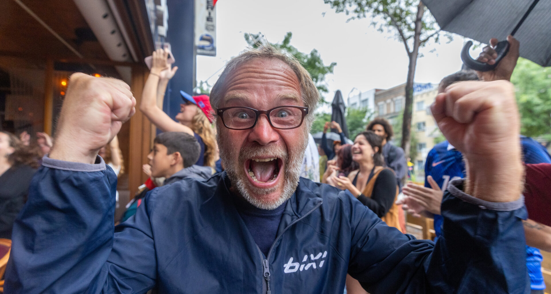 A man cheers in a very animated way toward the camera outside a bar.