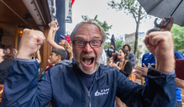 A man cheers in a very animated way toward the camera outside a bar.