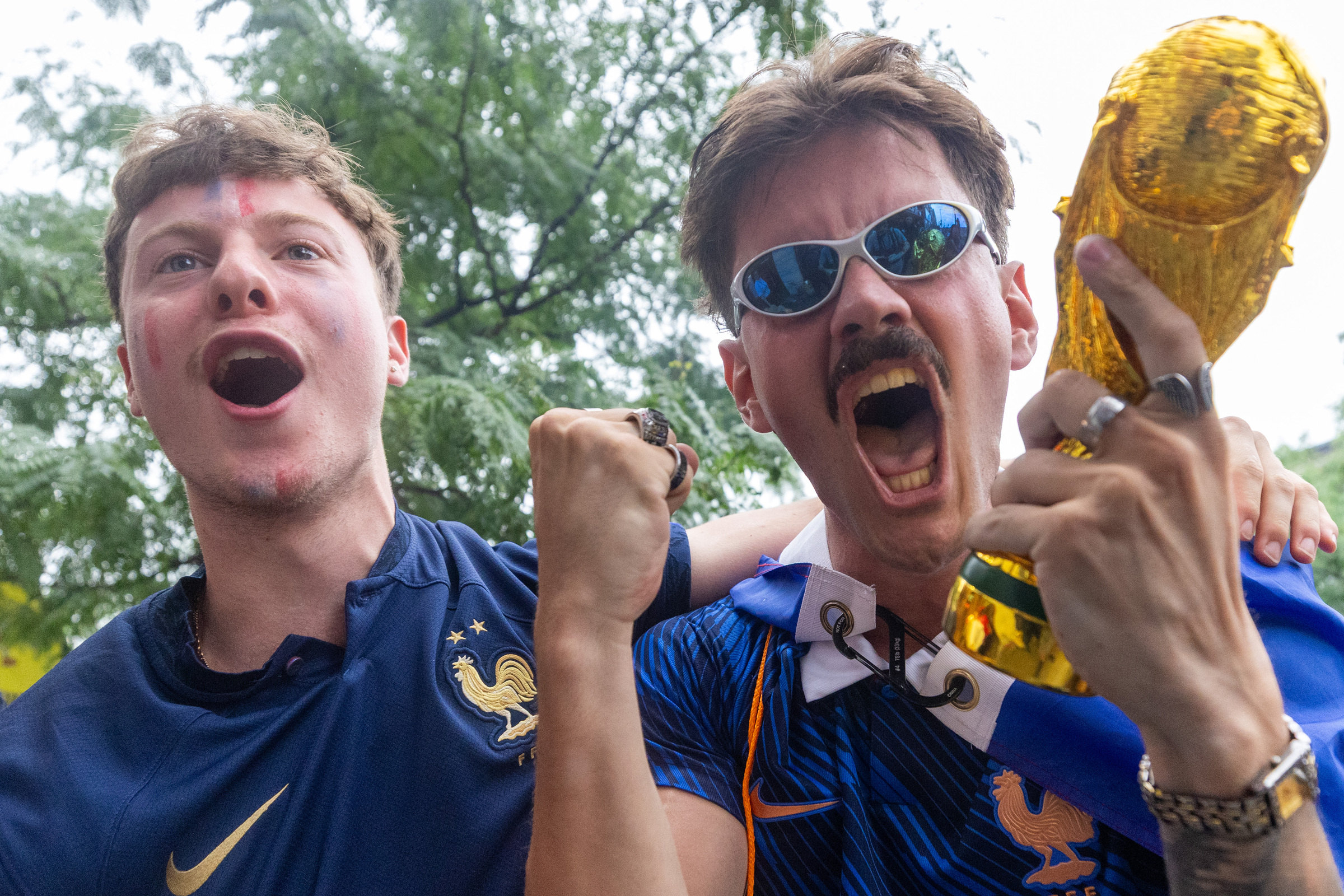 Two men dressed in blue cheer, one holding a World Cup trophy replica, outside.