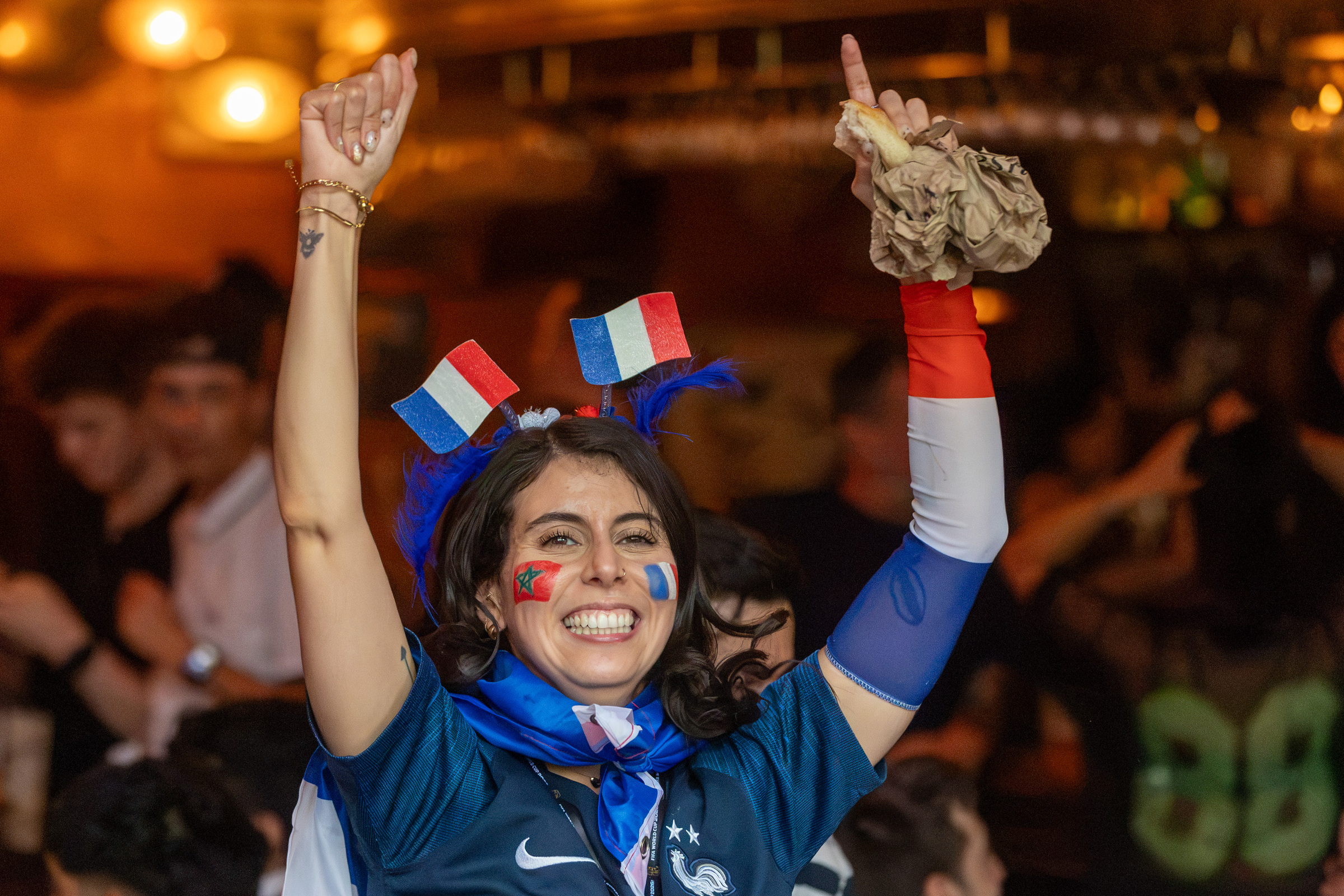 A woman with two French flags in her hair cheers in a bar.