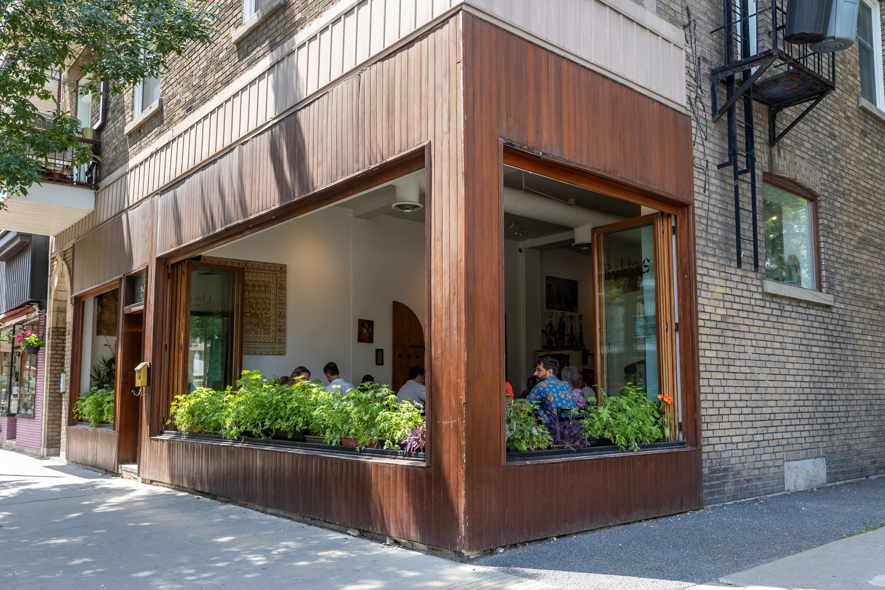 A restaurant with its windows open and flower boxes filled to the brim is seen on a street corner.