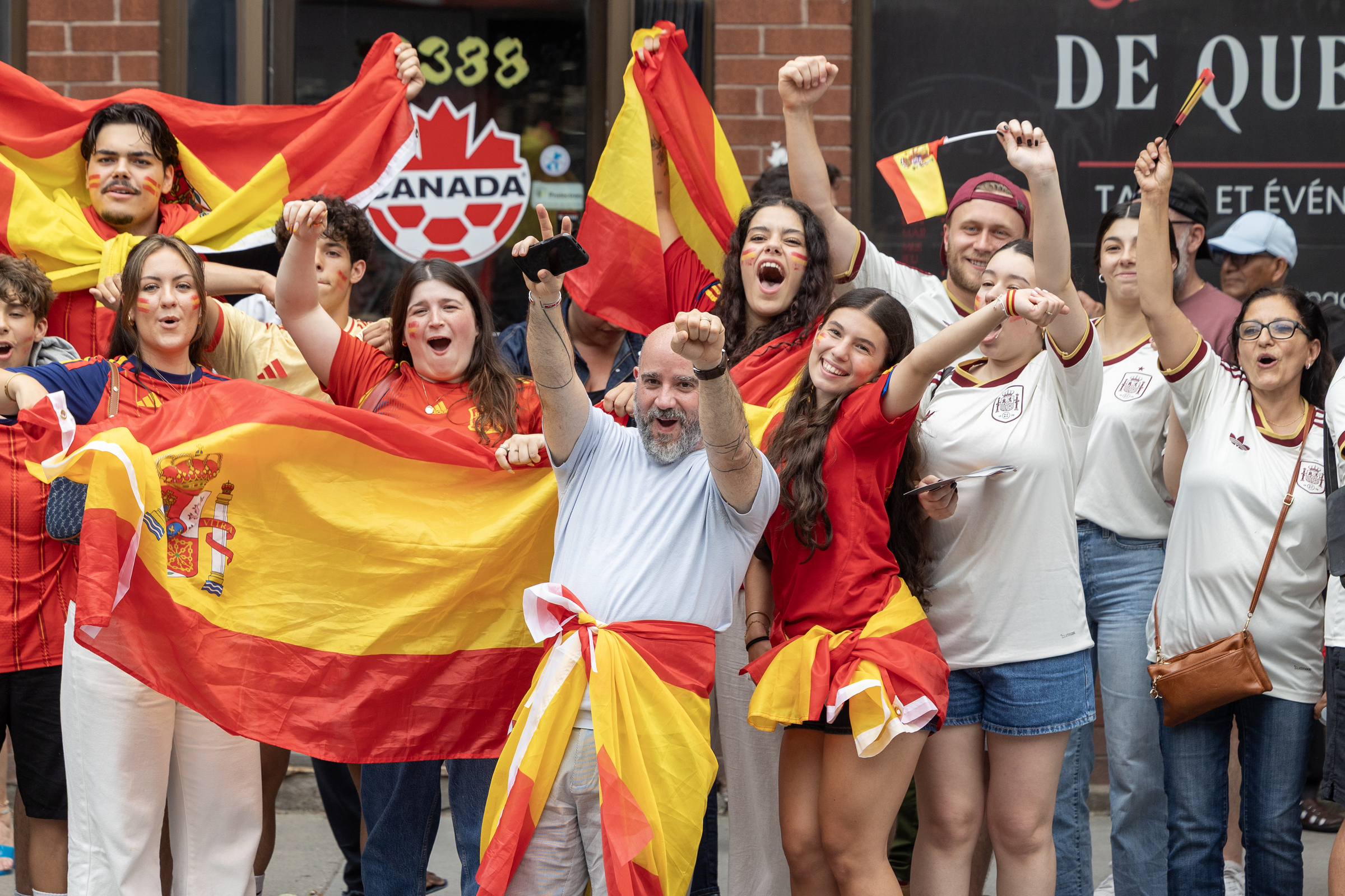 People wave Spanish flags lined up on a sidewalk outside.