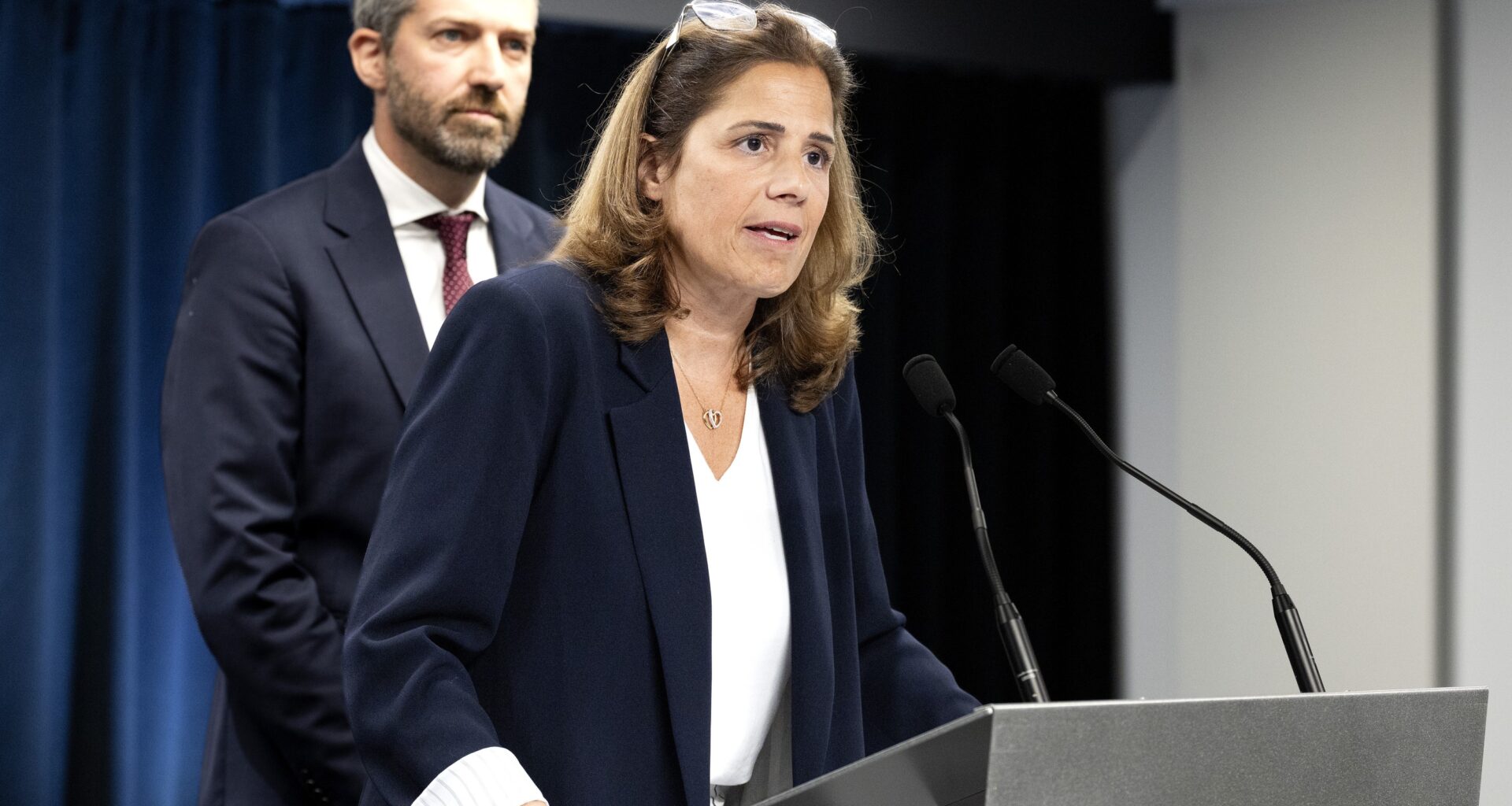 Woman at podium speaks into mic while man looks on behind her.
