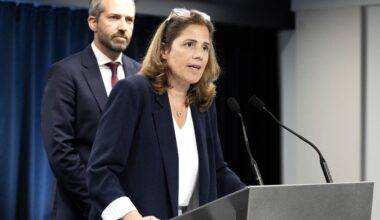 Woman at podium speaks into mic while man looks on behind her.