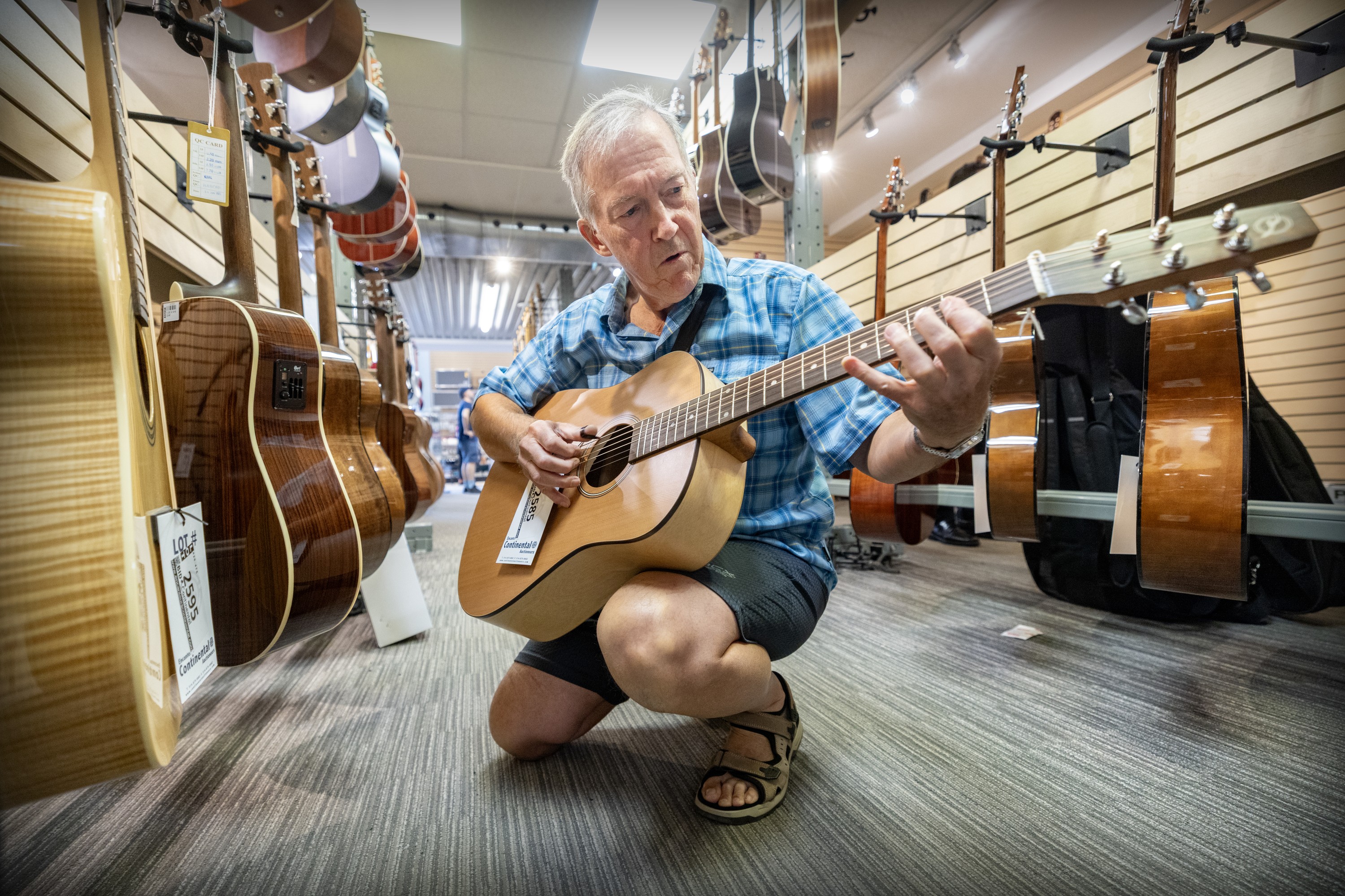 A man plays an acoustic guitar between rows of similar instruments in a music store