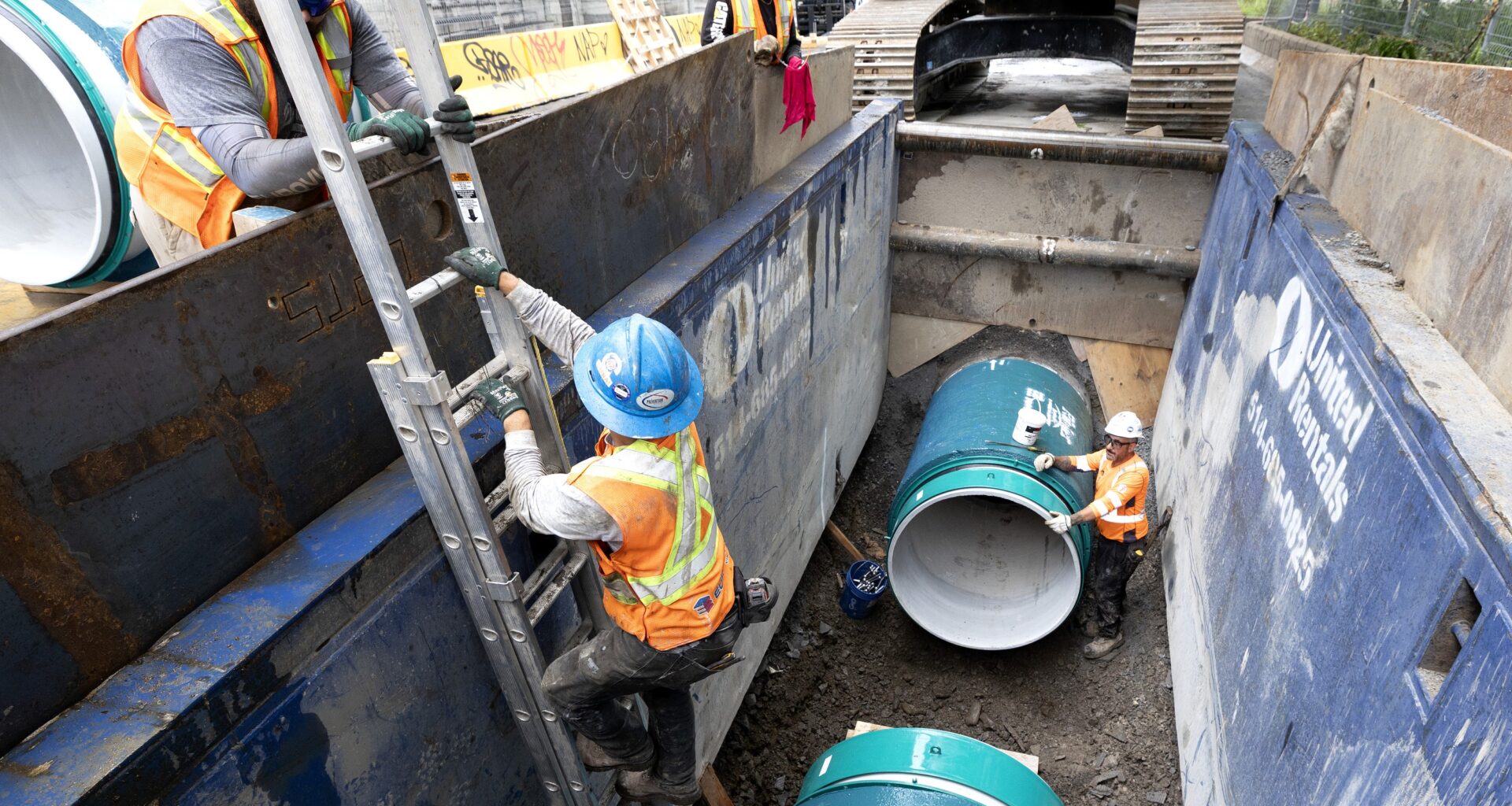 Workers in trench with big water main pipes.