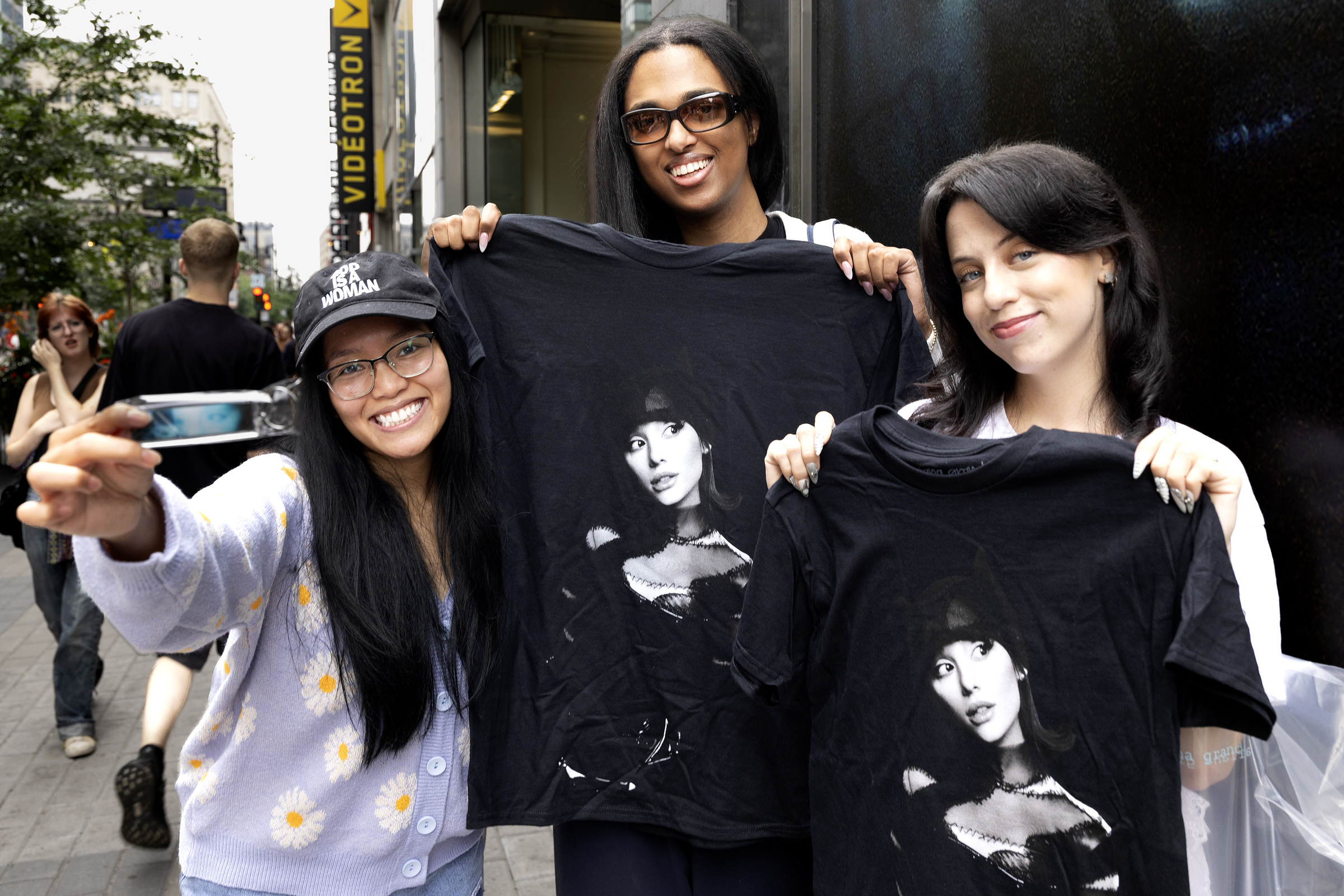 Two young women hold up black T-shirts with Ariana Grande's photo on them while a third holds up a keychain.