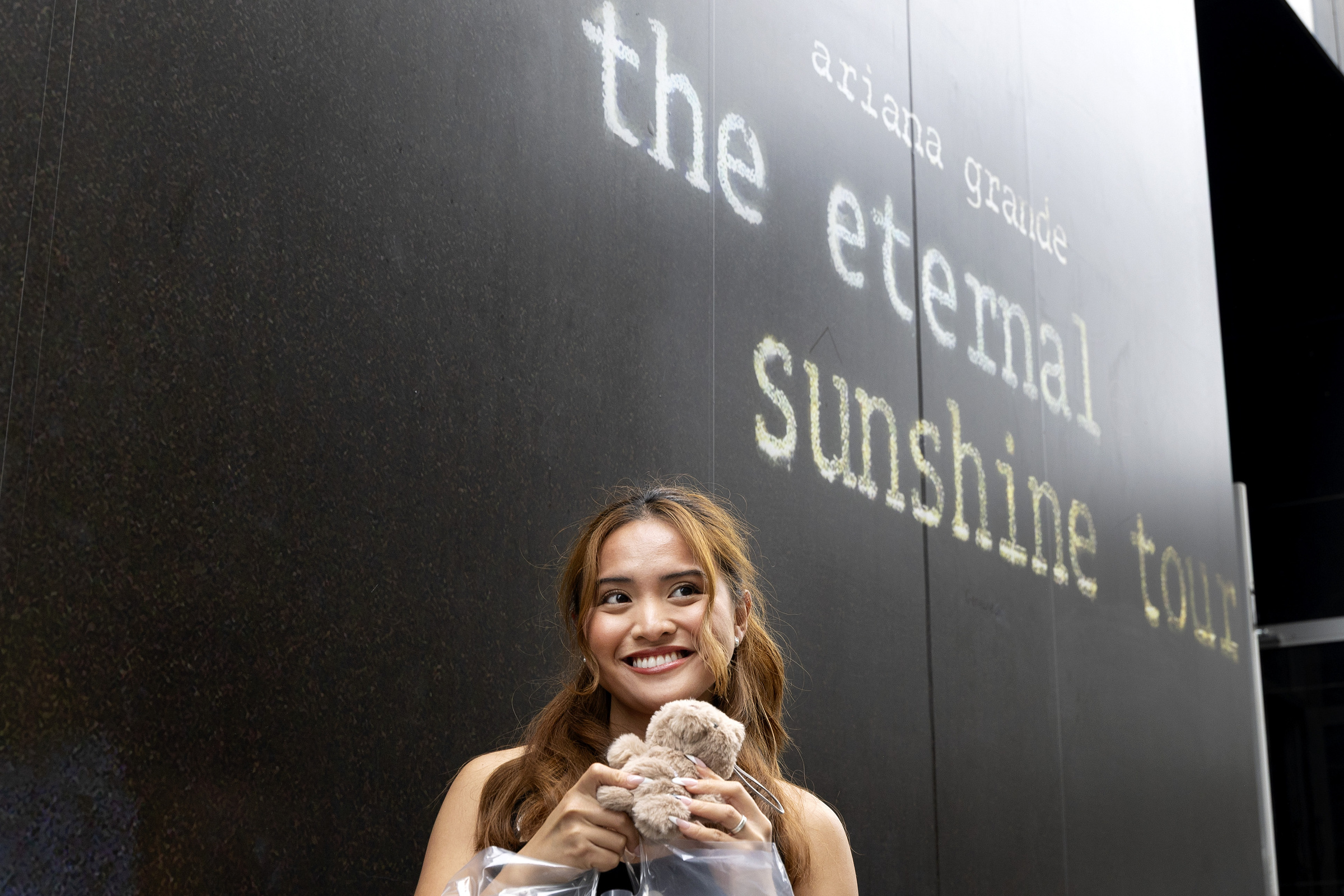 A young woman smiles as she holds a teddy bear in front of a large poster for Ariana Grande's The Eternal Sunshine tour.