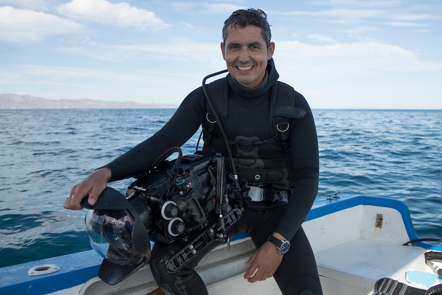 A man in a black scuba suit sitting on edge of a white boat with blue trim in the ocean, a massive underwater camera in his lap.