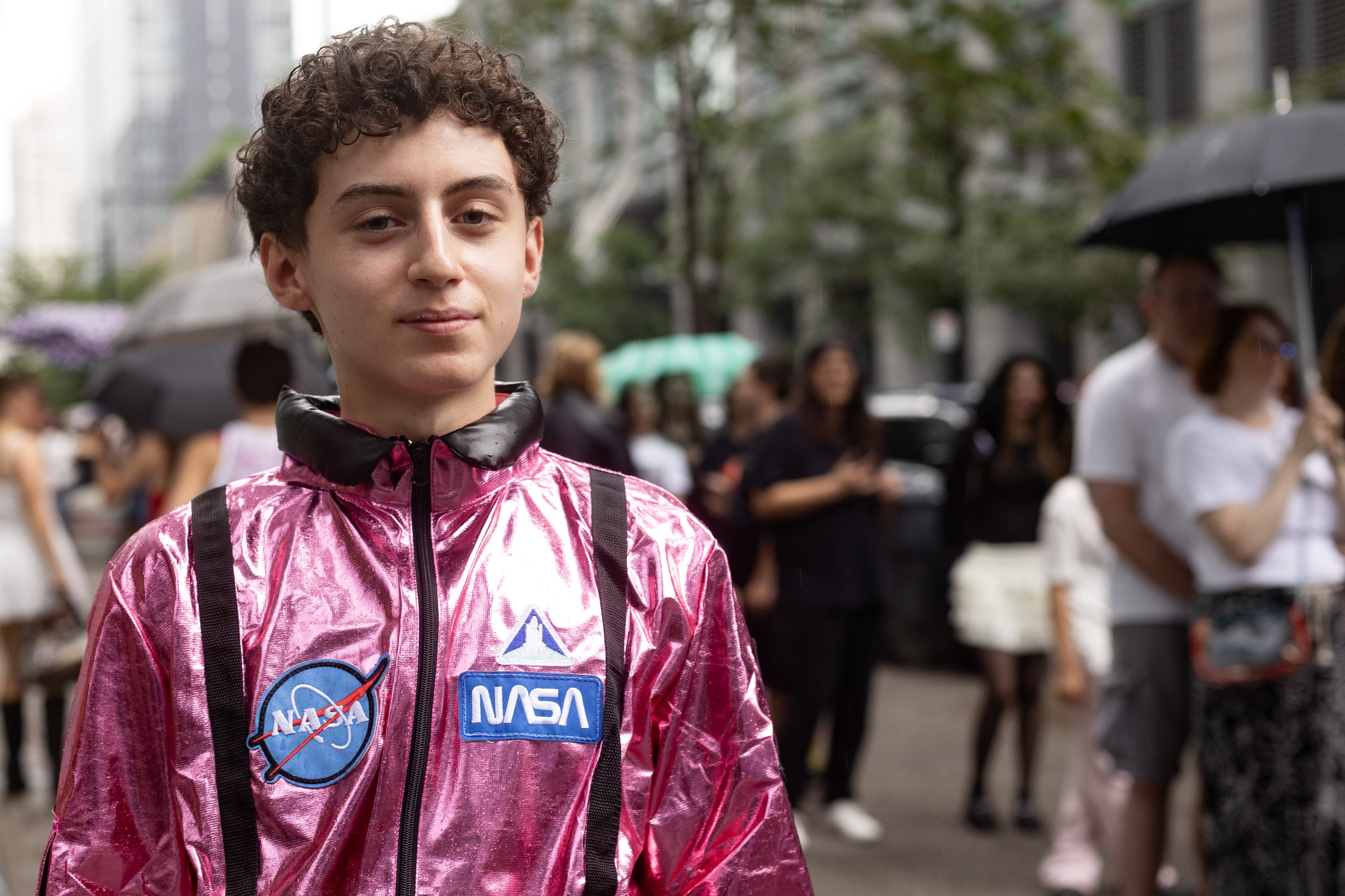 A young man in a shiny pink suit with NASA logos on it stands on a rainy sidewalk