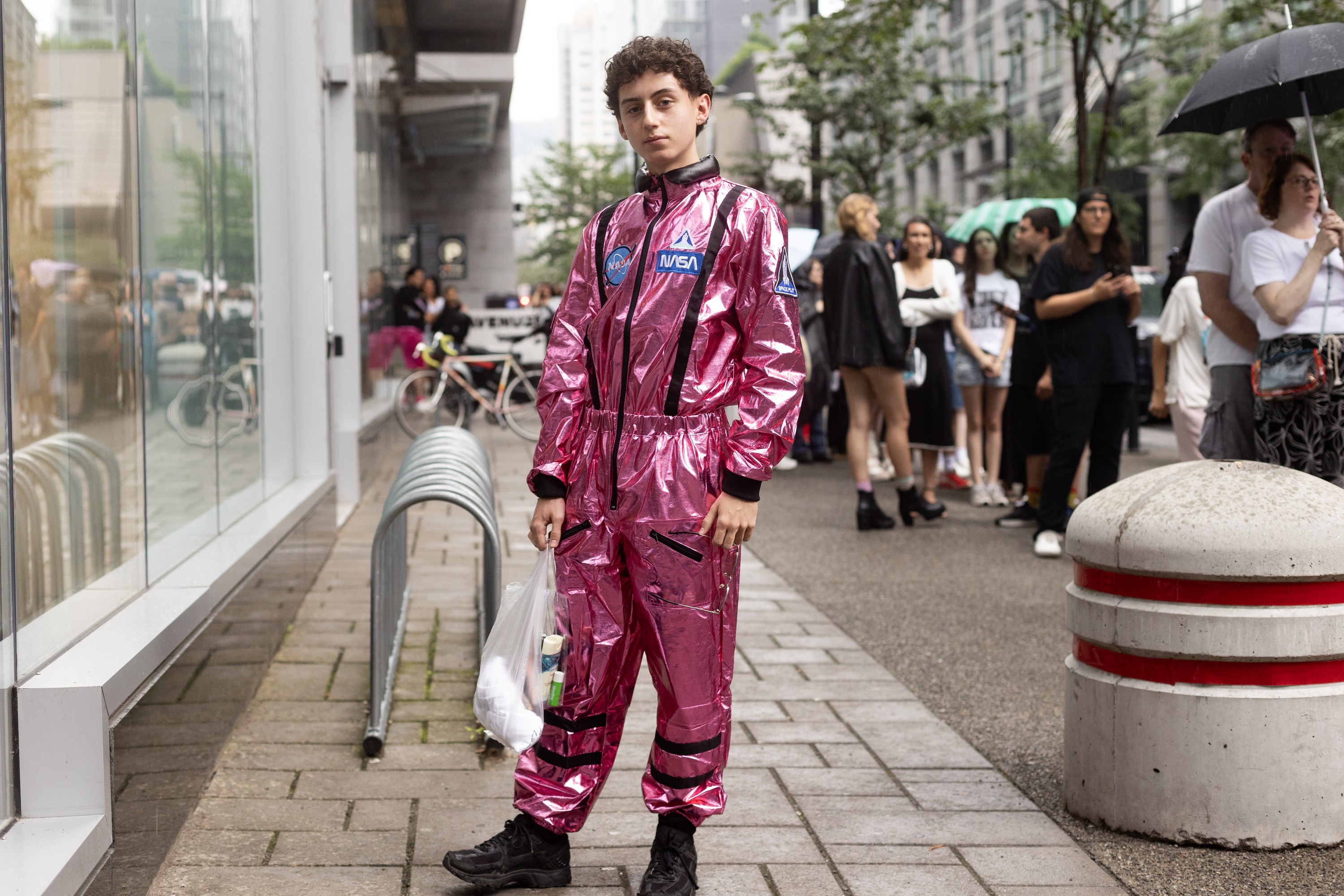 A young man in a shiny pink suit with NASA logos on it stands on a rainy sidewalk