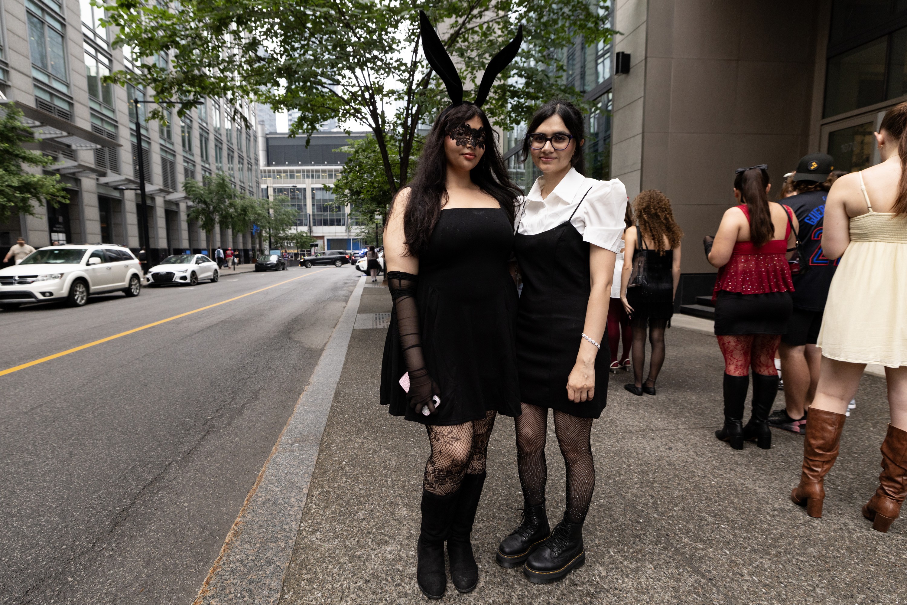 Two young women, one of them wearing an eye mask and large fake animal ears, stand on a sidewalk next to a line of people