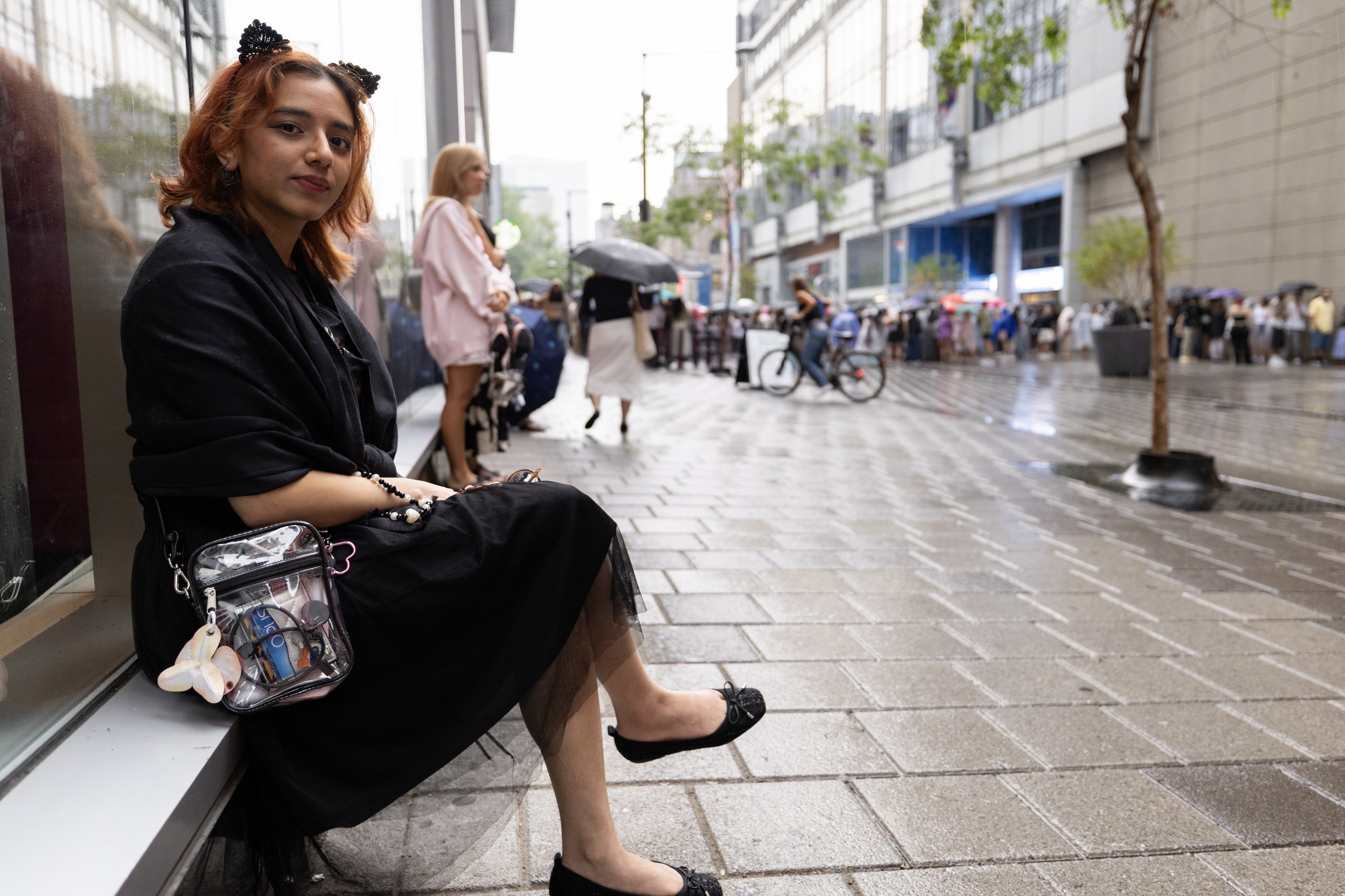 A person wearing a cat-ear headpiece sits on a ledge outside a building on a rainy street