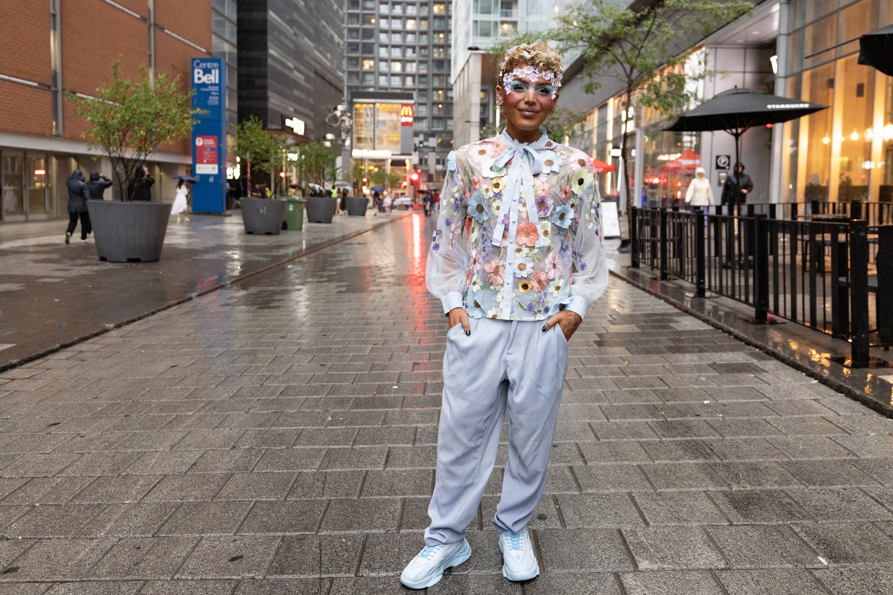 A person in colourful floral appliqués and a floral top looks at the camera while standing on a rainy street