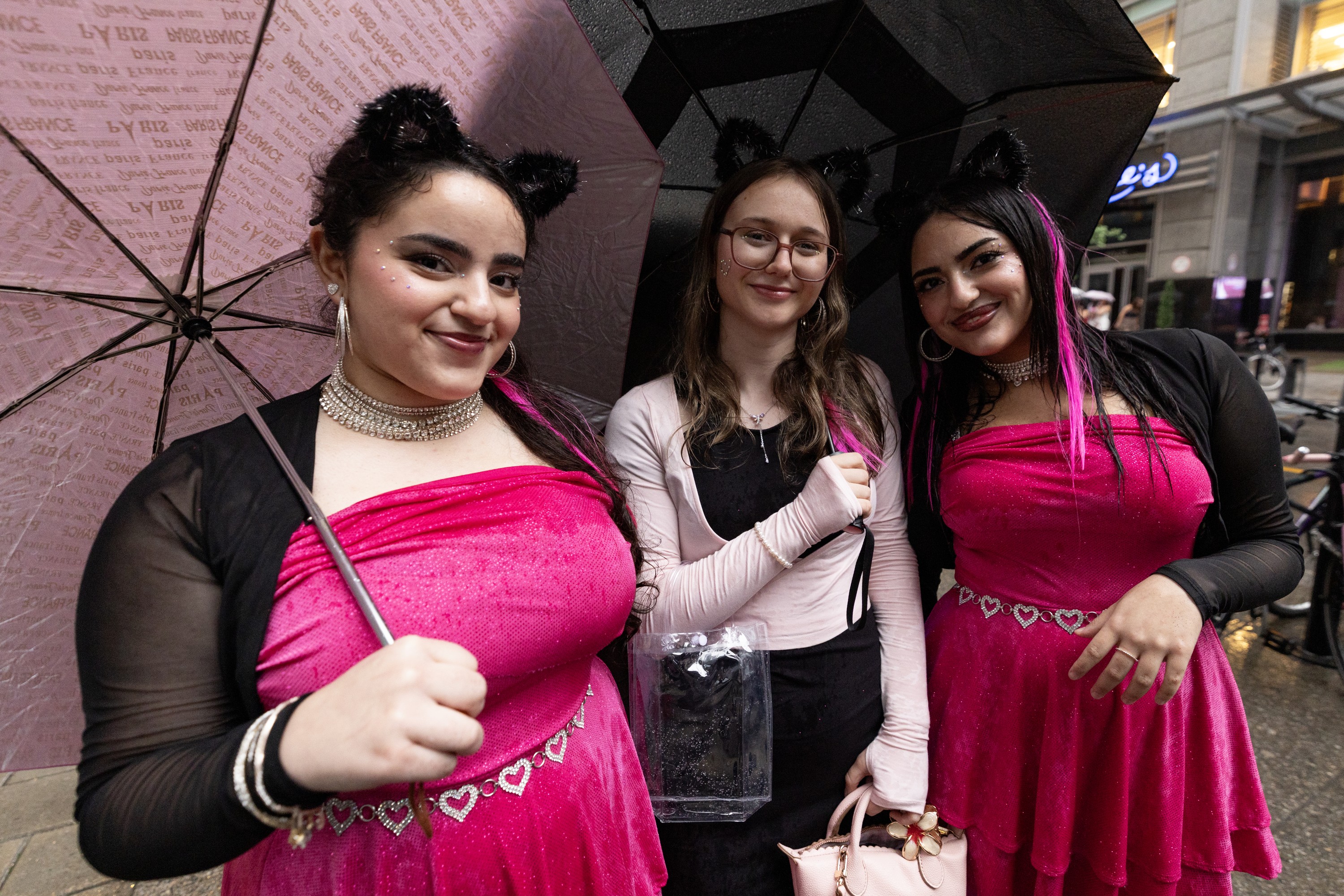 Three young women wearing cat-ear headpieces congregate under umbrellas on a rainy street