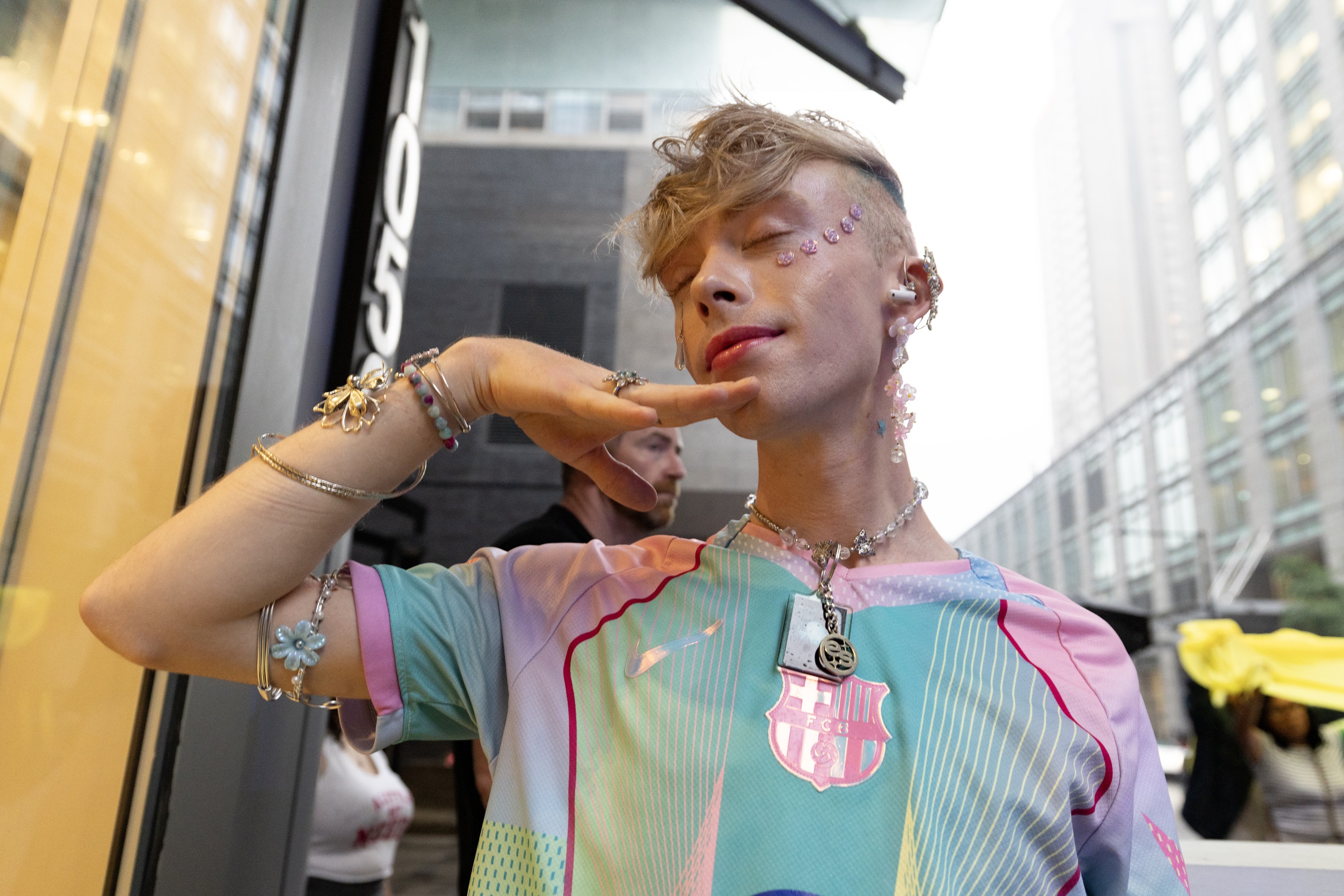 A person wearing jewelry and accessories holds their arm up to their face as they close their eyes while standing on a street