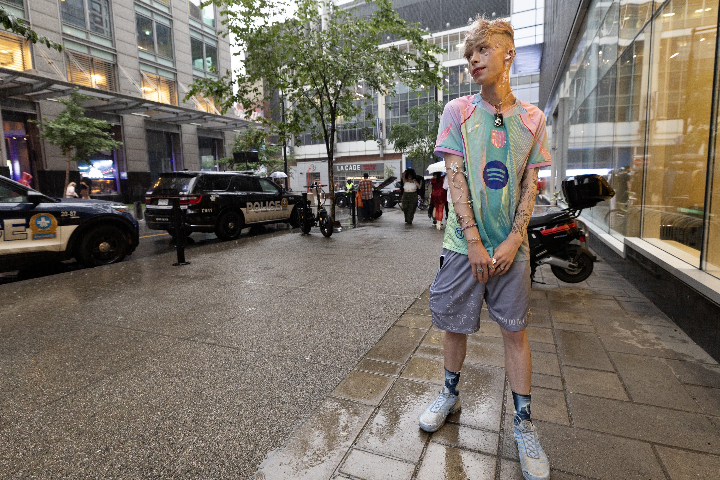 A person with stylish hair and accessories stands on a rainy street, looking to the side