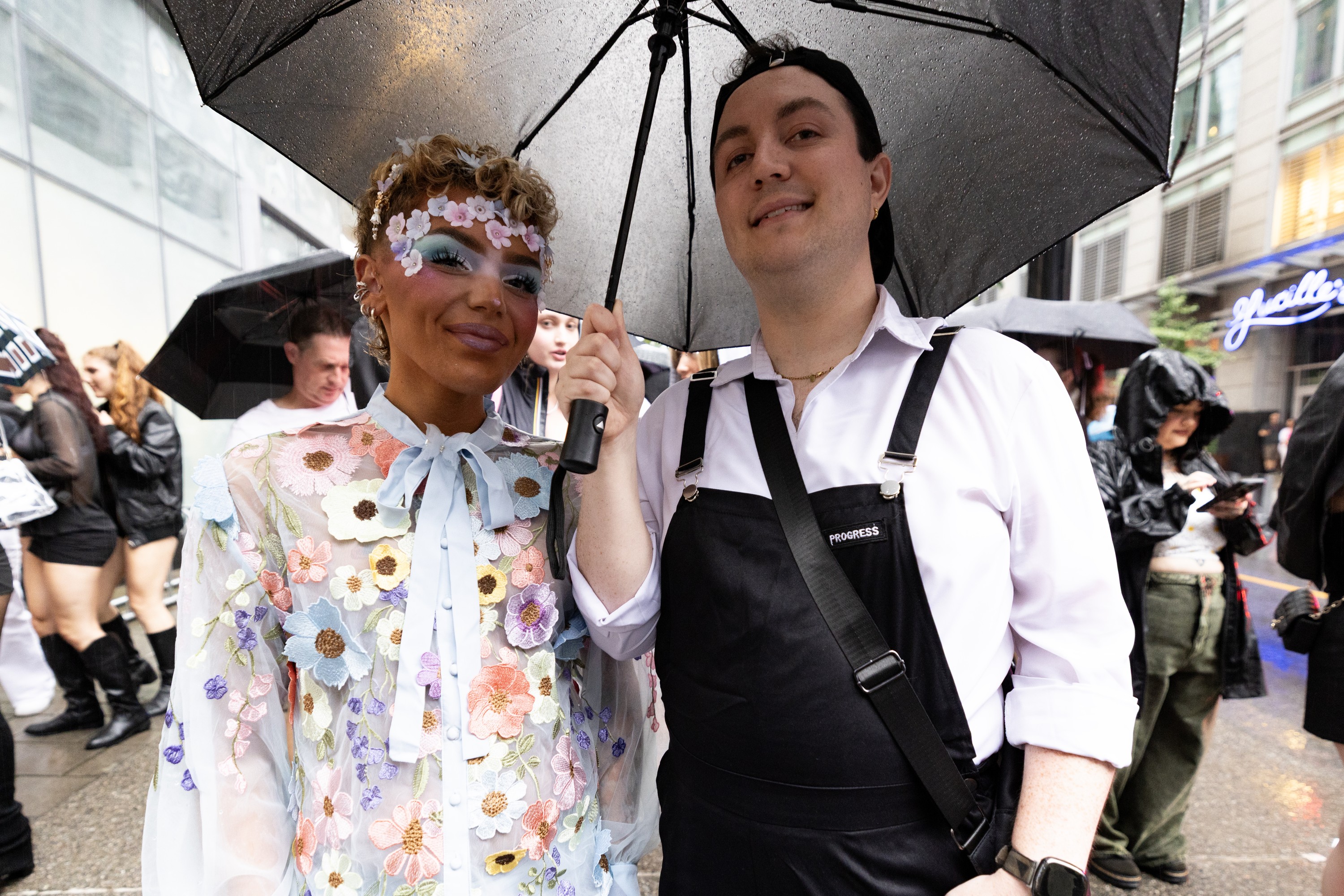 Two people – one in overalls, the other in floral appliqués and a floral top – are under an umbrella on a busy street